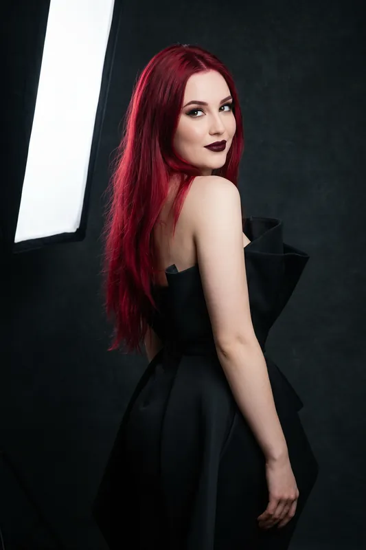 Donna con lunghi capelli cremisi, in un abito nero strutturato, che guarda da sopra la spalla in uno studio scuro.