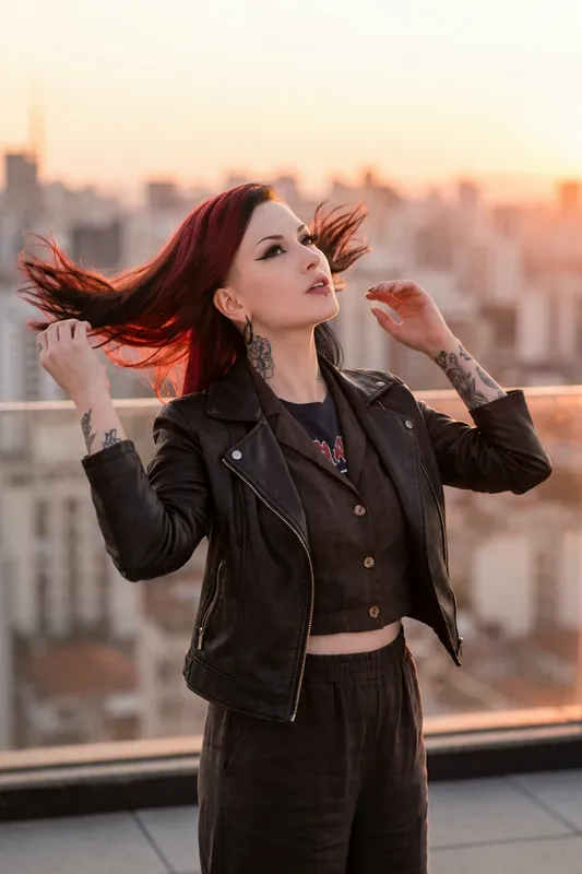 Donna con capelli rossi e neri e braccia tatuate, in giacca di pelle, mentre scuote i capelli su un tetto al tramonto.